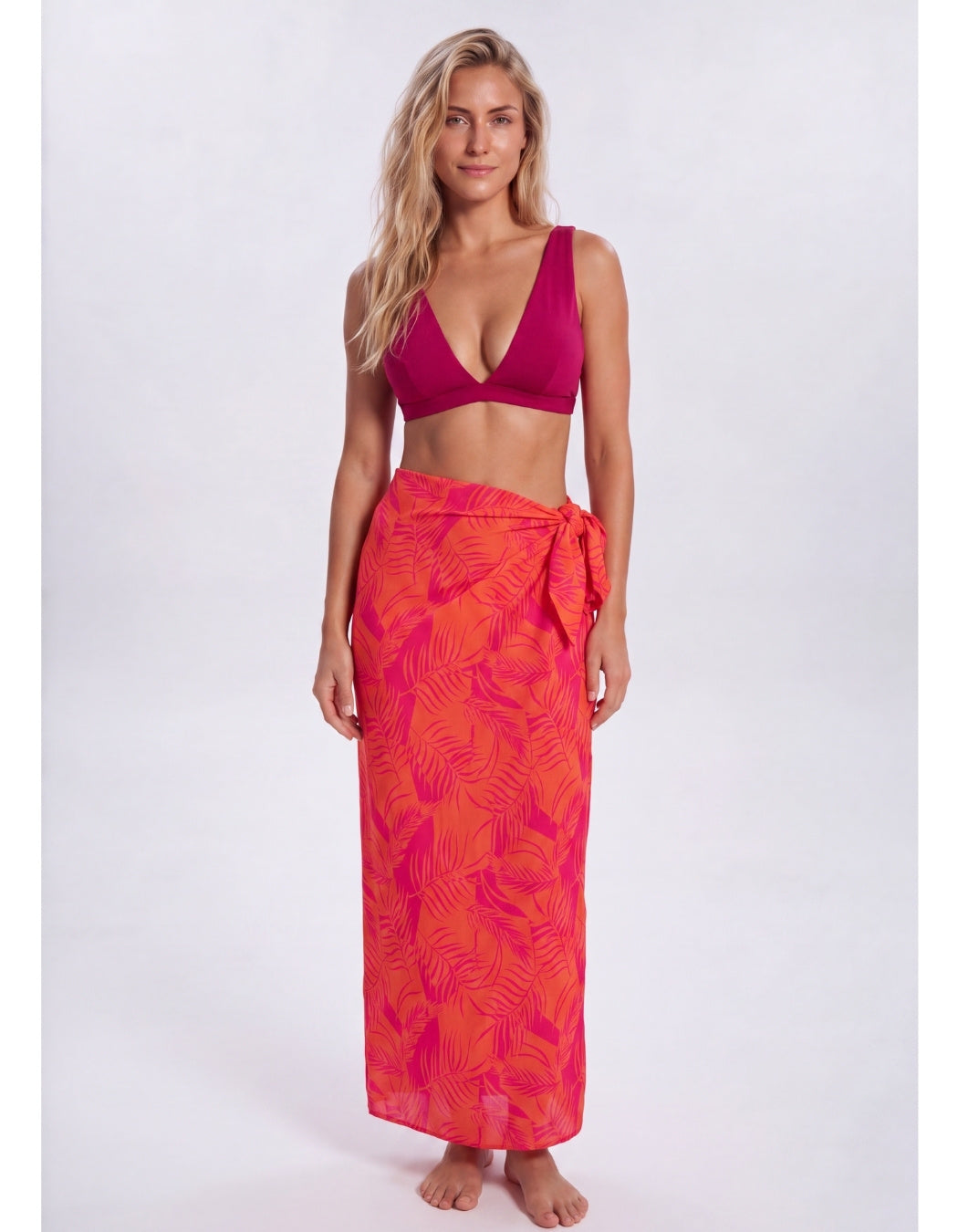 A woman modeling a two-piece swimwear set consisting of a deep magenta pink bikini top and a matching orange and pink tropical leaf print sarong wrap skirt, shot against a plain light gray studio background. Sandbar Swimwear