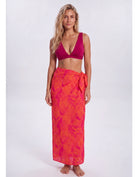 A woman modeling a two-piece swimwear set consisting of a deep magenta pink bikini top and a matching orange and pink tropical leaf print sarong wrap skirt, shot against a plain light gray studio background. Sandbar Swimwear