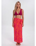 A woman modeling a two-piece swimwear set consisting of a deep magenta-burgundy bikini top and a bright pink-orange tropical leaf print sarong wrap skirt, shot against a seamless light gray studio background. Sandbar Swimwear