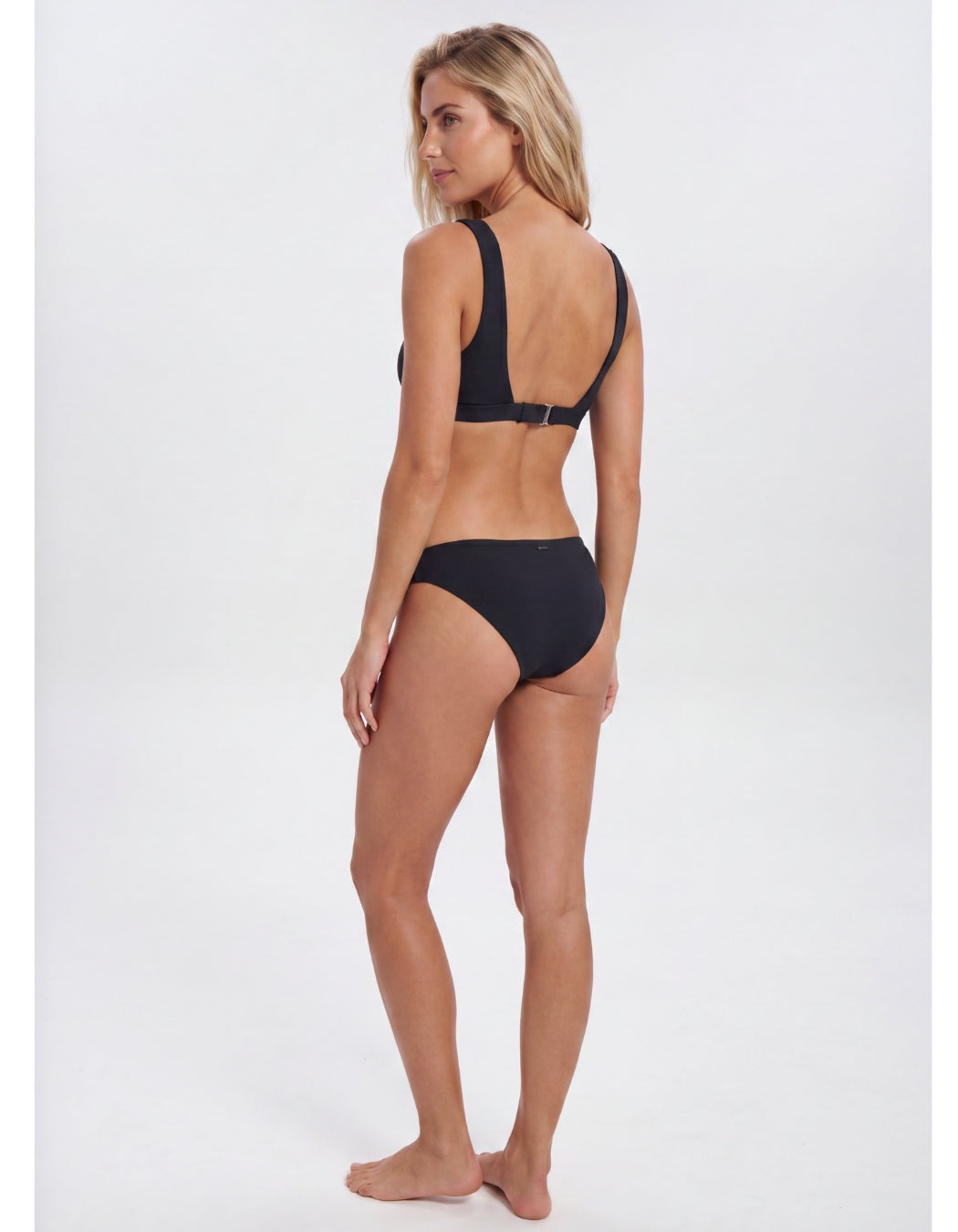 A woman modeling a black two-piece bikini from the back, standing against a plain light gray studio background. Sandbar Swimwear