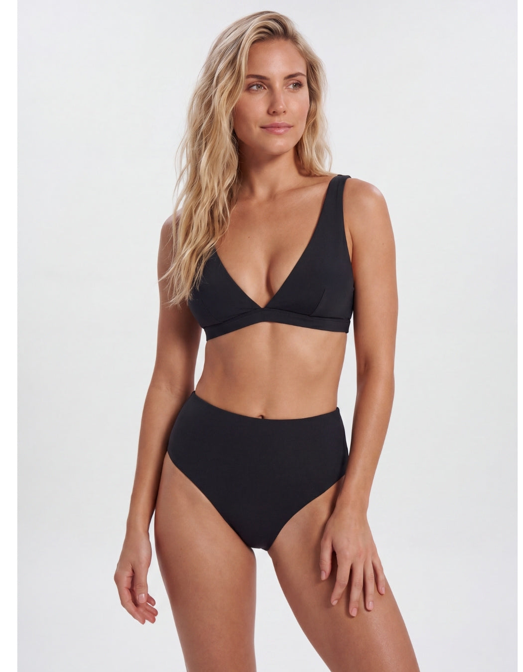A blonde model wearing a black two-piece bikini with a deep V-neck triangle top and high-waisted bottoms, photographed against a light gray studio background. Sandbar Swimwear