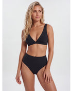 A blonde model wearing a black two-piece bikini with a deep V-neck triangle top and high-waisted bottoms, photographed against a light gray studio background. Sandbar Swimwear