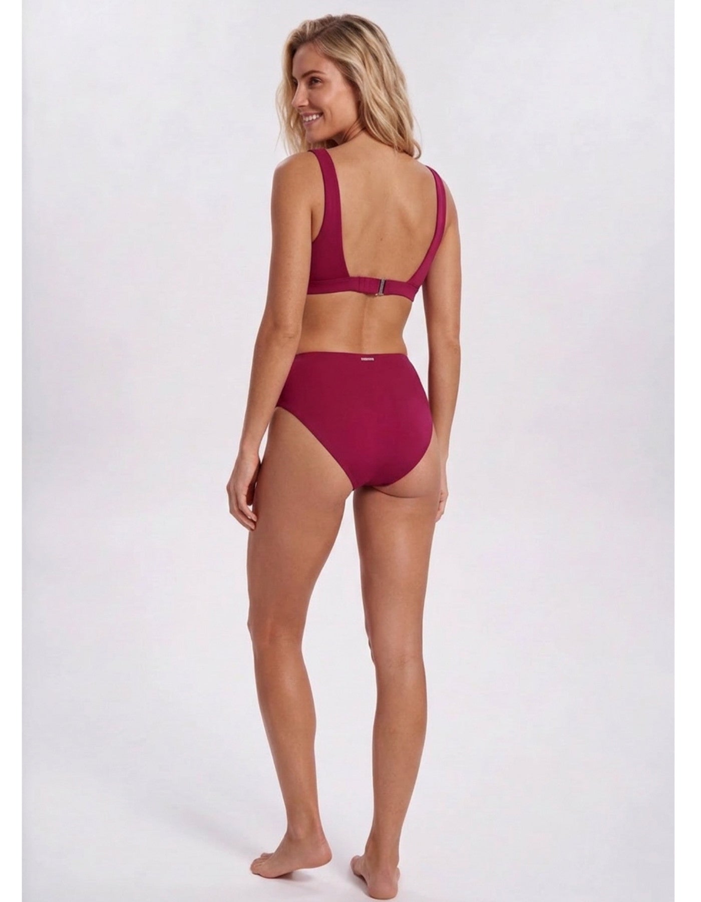 A woman modeling a two-piece bikini from the back, featuring a deep scoop neck and high-waisted bottoms, in a solid deep magenta color, set against a plain light gray studio background. Sandbar Swimwear