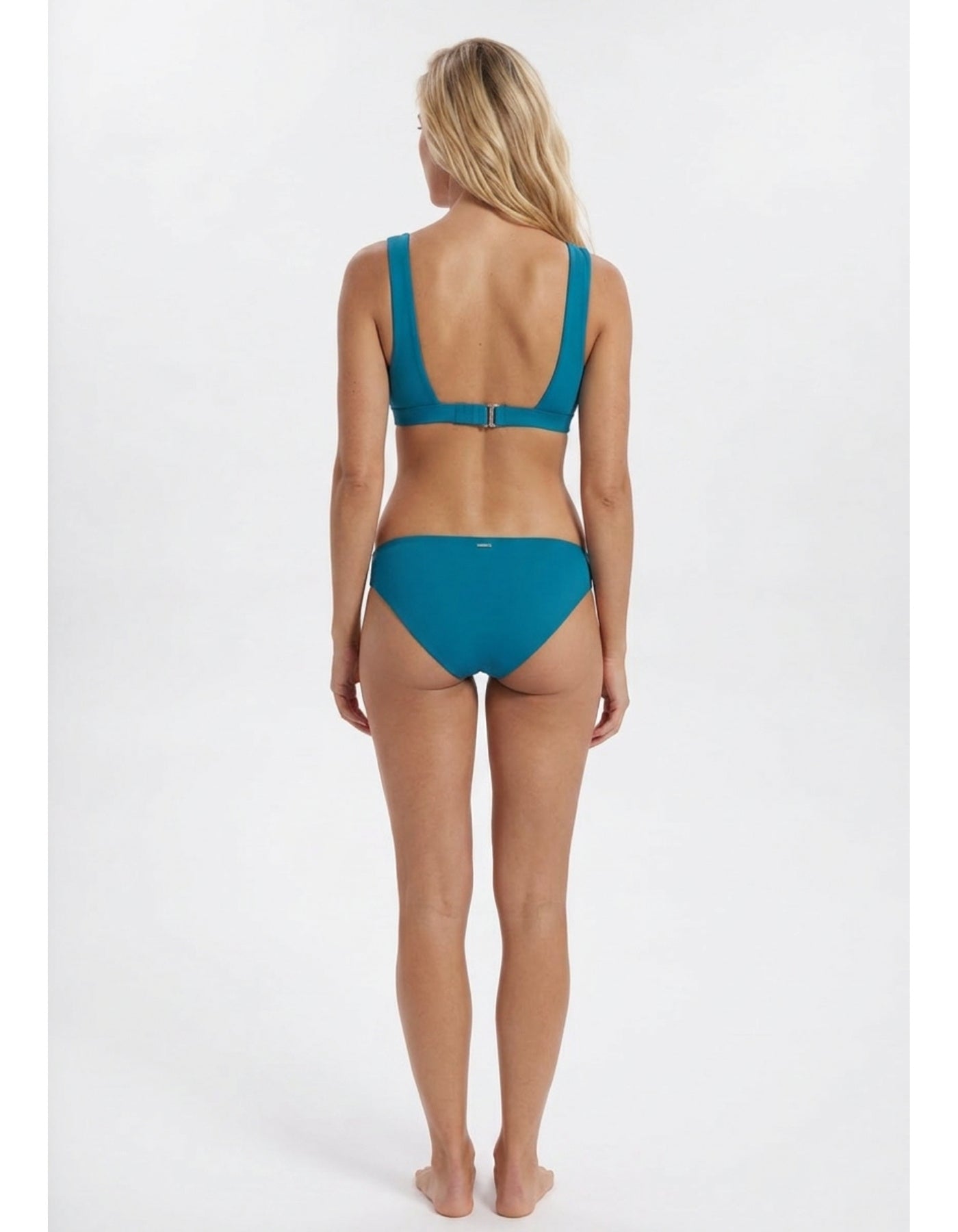 A woman viewed from the back wearing a teal two-piece bikini against a plain light gray studio background Sandbar Sustainable Swimwear