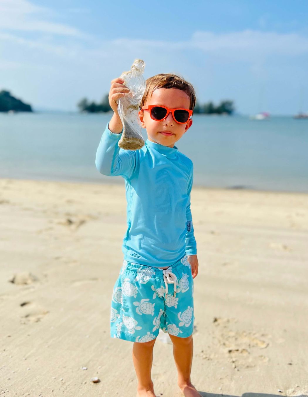 Boy on the beach in Sandbar swimwear holding up a plastic bottle