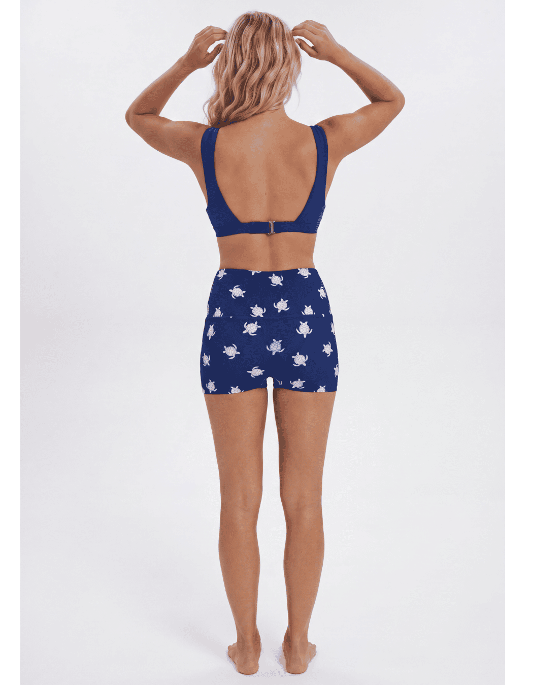 A woman shown from the back wearing a two-piece swimsuit with a solid navy blue top and matching high-waisted shorts featuring a white floral pattern, set against a plain light gray background. Sandbar Swimwear