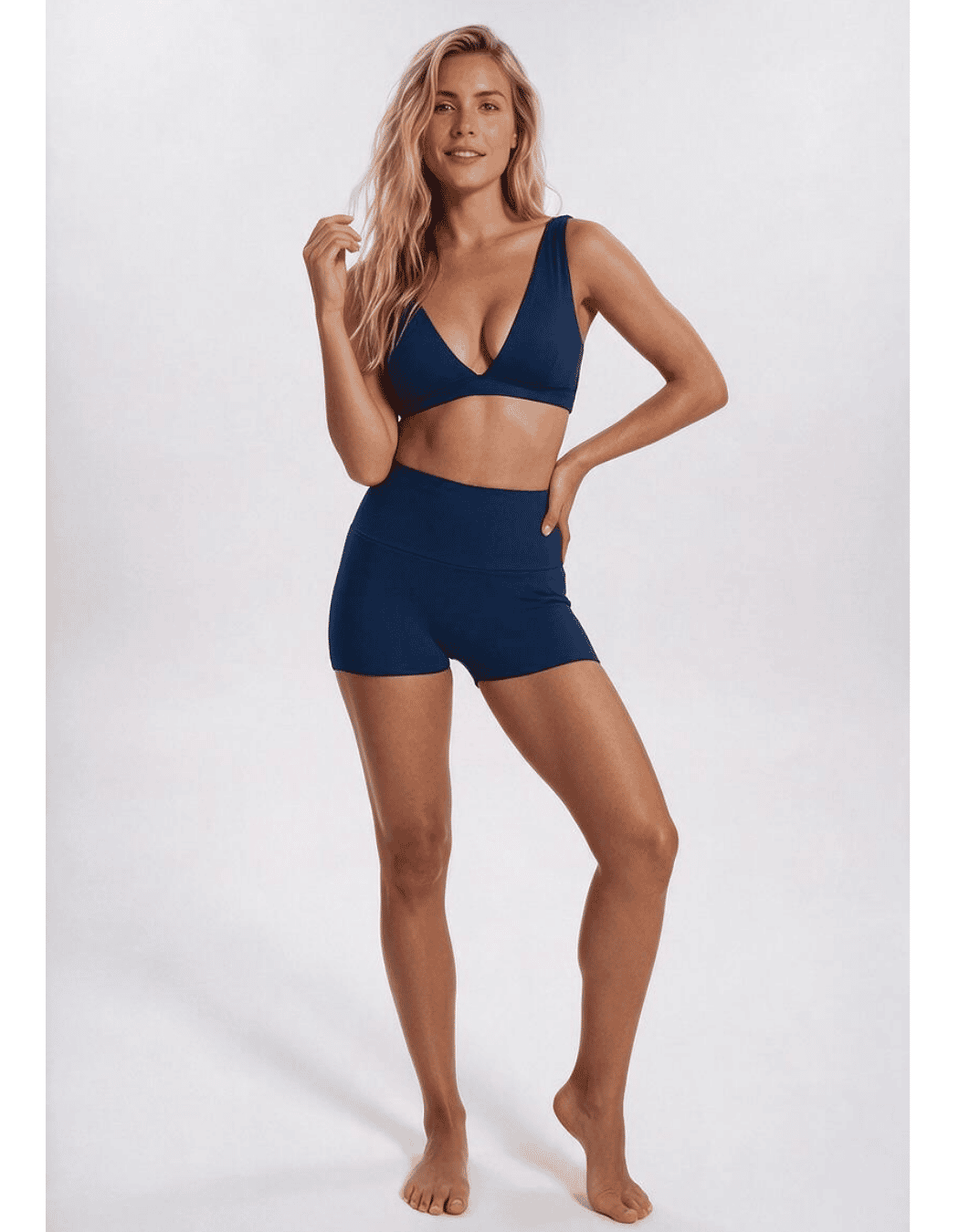 A woman modeling a matching two-piece swimwear set consisting of a navy blue triangle bikini top and high-waisted navy blue bikini shorts, posed against a light gray studio background. Sandbar Swimwear