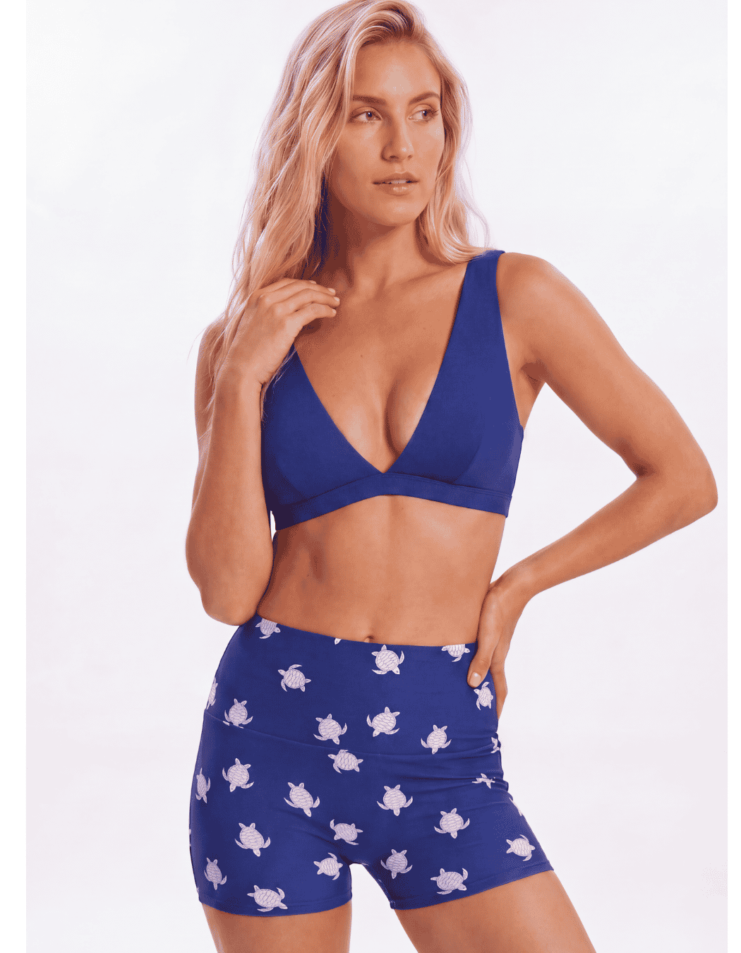 A woman modeling a royal blue two-piece swimsuit with a white turtle print, consisting of a triangle bikini top and matching high-waisted shorts, against a plain white background Sandbar Swimwear
