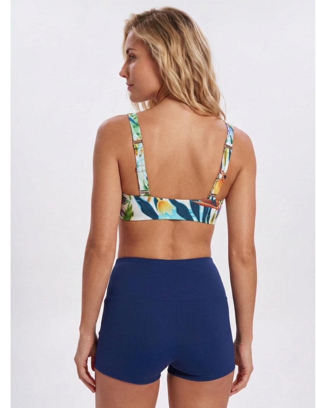 A woman shown from the back wearing a two-piece swimsuit with a tropical floral print bikini top and solid navy blue high-waisted shorts, against a light gray studio background. Sandbar Swimwear