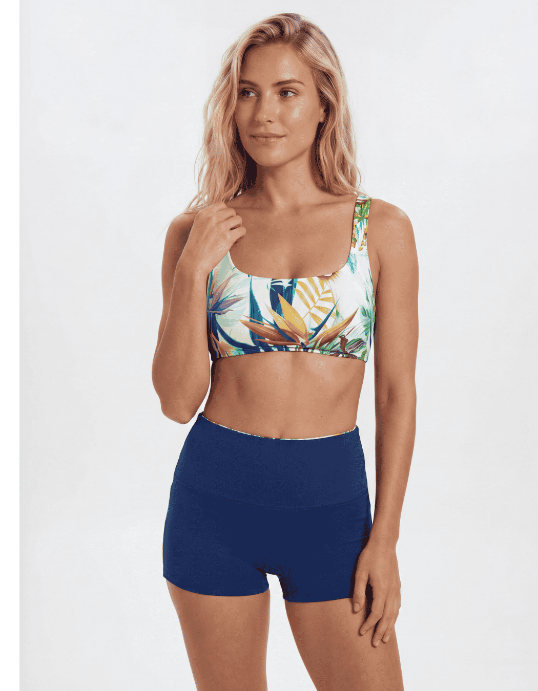 A woman modeling a two-piece swimwear set featuring a tropical print sports bra and solid navy blue high-waisted shorts, set against a plain light gray background Sandbar Swimwear