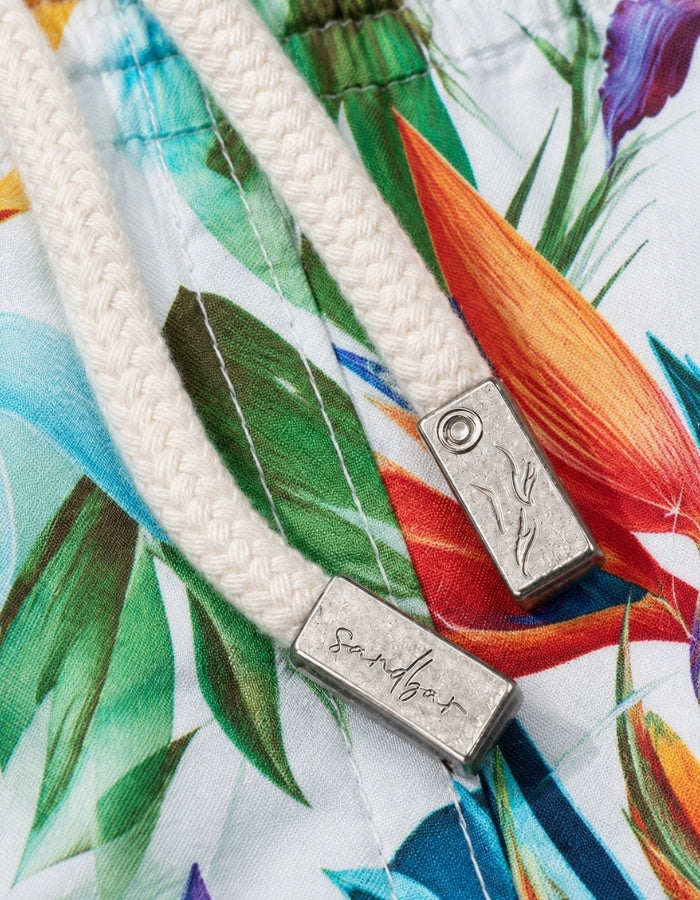 Close-up of a white braided drawstring with a silver metal toggle engraved with the brand name 'Sandbar', resting on a white fabric background with a colorful tropical floral print. Sandbar Swimwear shorts detail
