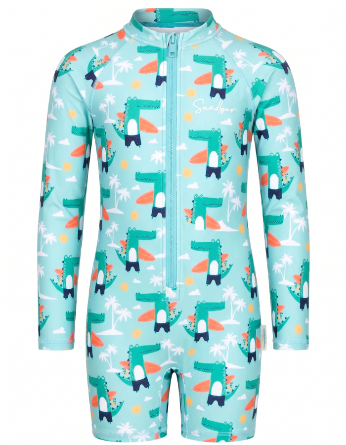 A children's long-sleeve one-piece swimsuit or sun protection suit in light aqua, featuring a repeating cartoon dinosaur pattern with surfboards, palm trees, and small yellow circles, plus a front zipper.