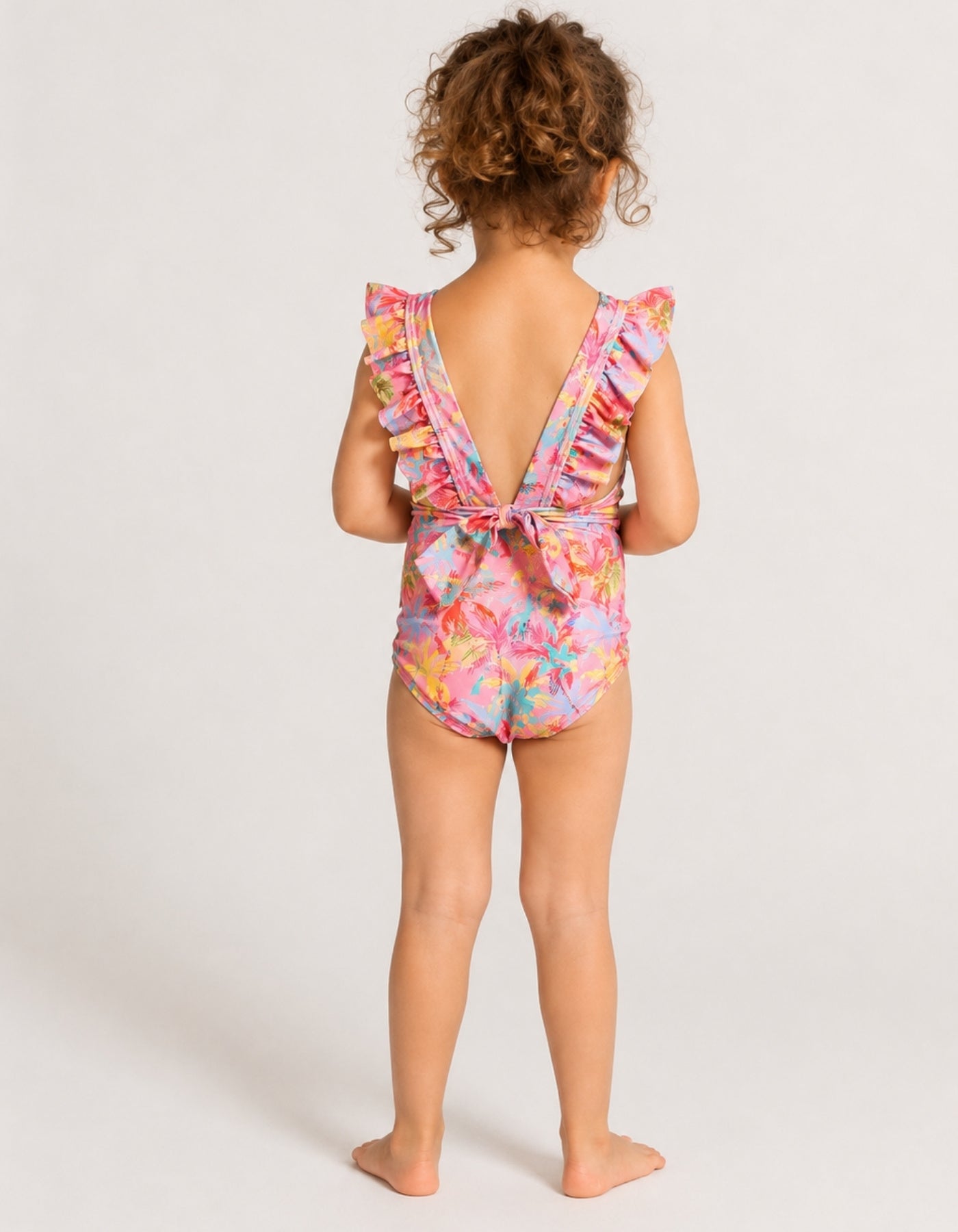 A young child viewed from behind wearing a colorful floral one-piece swimsuit with ruffled shoulder straps and a tie-back detail, standing against a plain light gray studio background.
