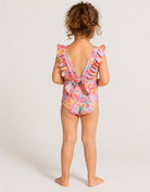 A young child viewed from behind wearing a colorful floral one-piece swimsuit with ruffled shoulder straps and a tie-back detail, standing against a plain light gray studio background.