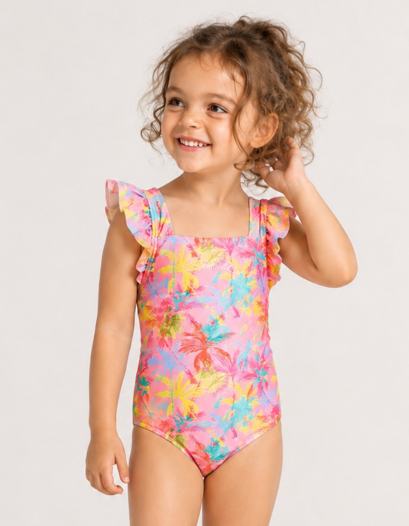 A young girl with curly brown hair wearing a vibrant pink one-piece swimsuit with a tropical multicolor floral print and ruffled shoulder straps, posing against a light gray studio background.