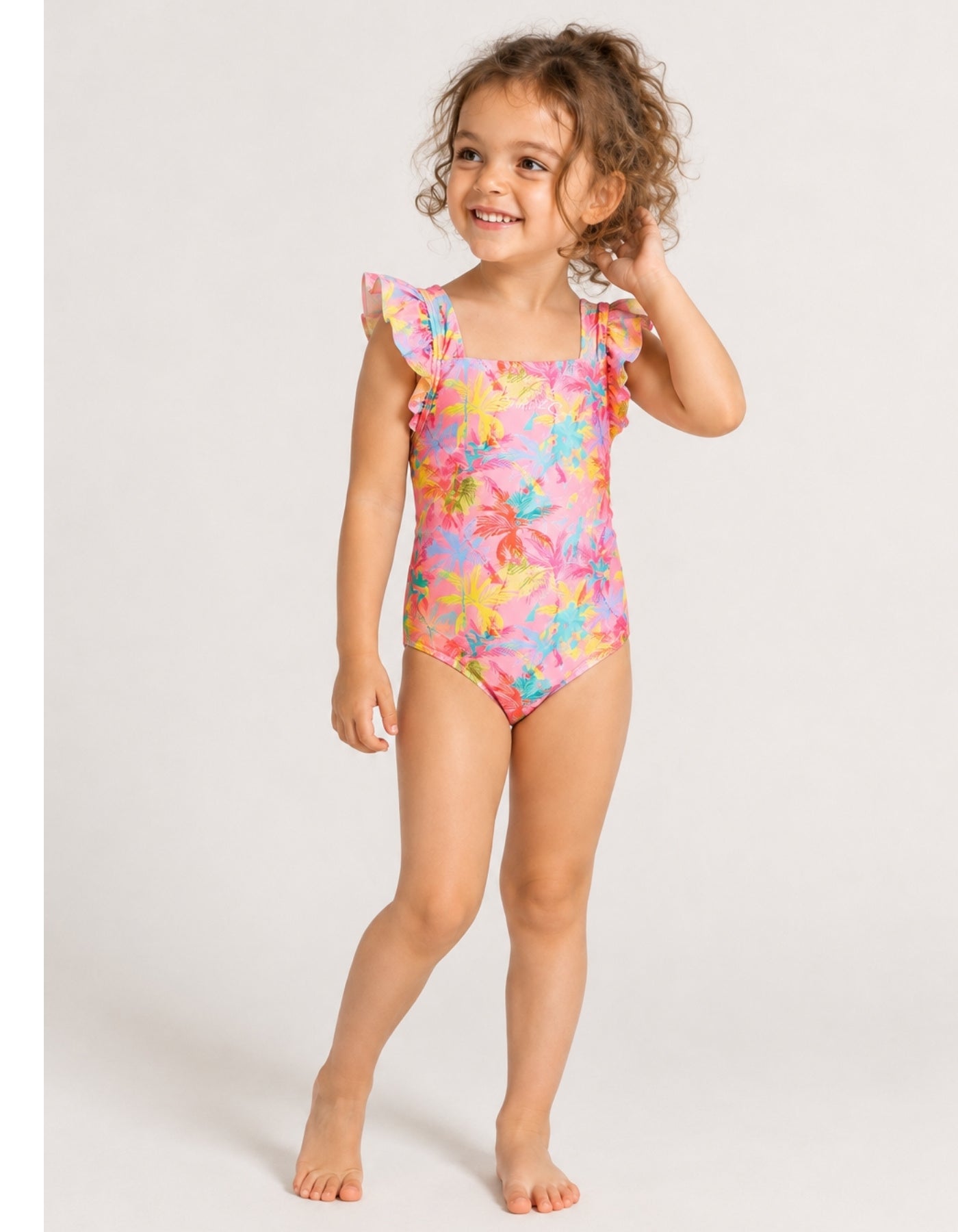 A young girl with curly brown hair stands barefoot against a light gray studio background, wearing a one-piece swimsuit with a bright multicolor floral print and ruffled shoulder straps.