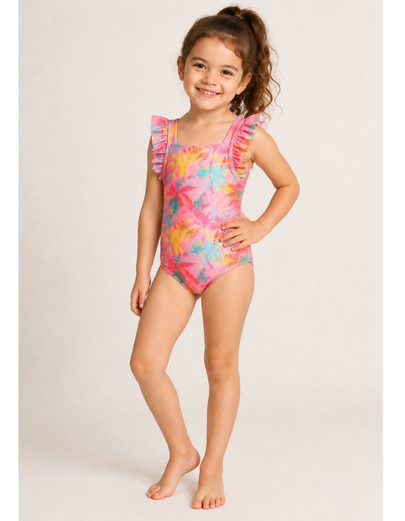 A young girl with curly brown hair stands barefoot against a light beige studio background, wearing a one-piece swimsuit with a bright pink base and a multicolor tropical floral print in yellow, blue, and orange. The swimsuit features ruffled shoulder straps.