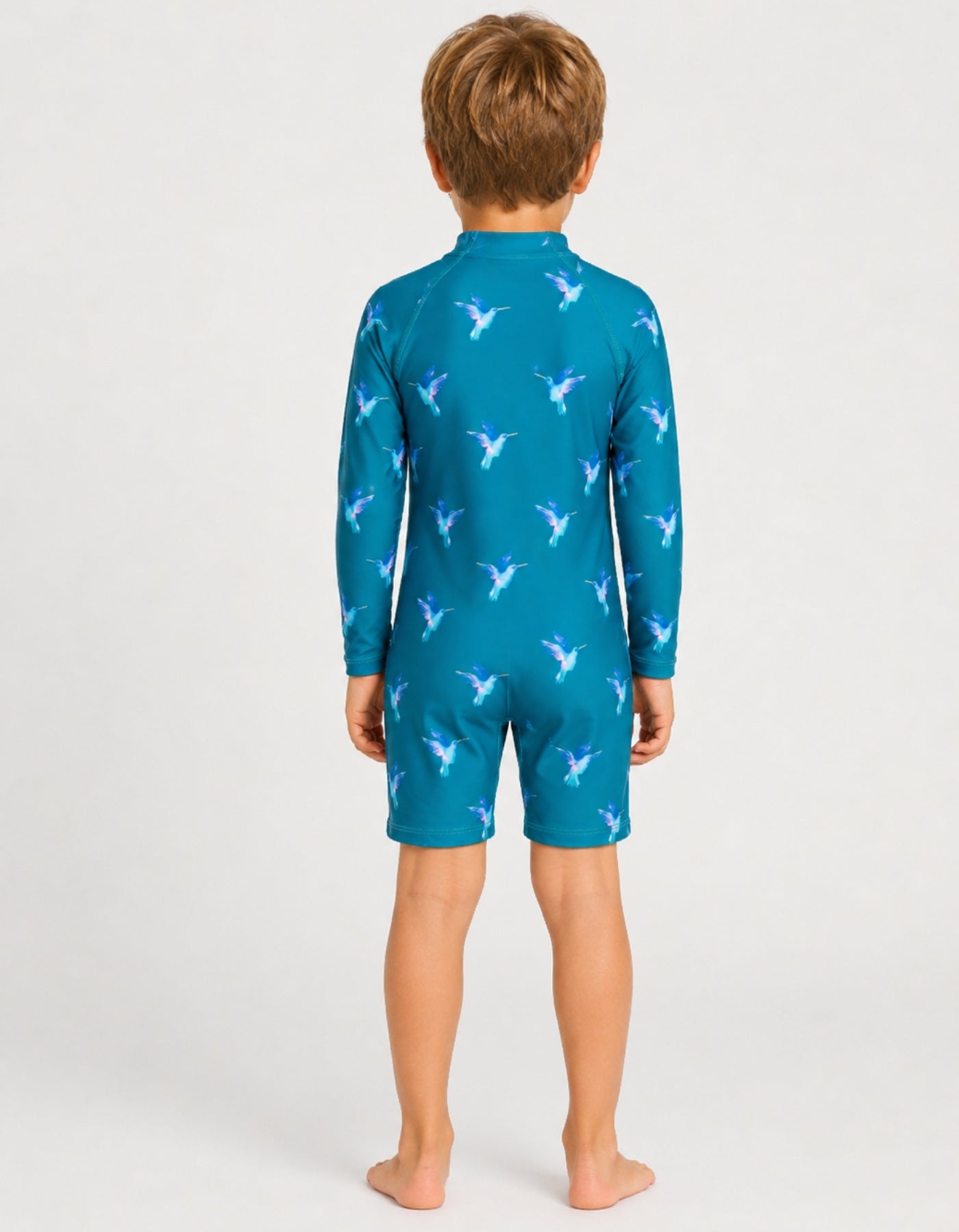 A child model viewed from the back wearing a teal long-sleeve swim shirt and matching swim shorts with a repeating white bird pattern, set against a plain light gray studio background.