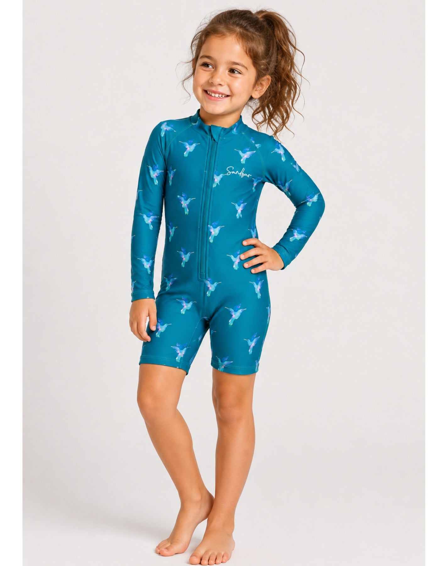 A young girl with her hair in a ponytail stands barefoot against a light gray studio background, wearing a teal-blue full-body swim coverall with a white allover bird pattern and a front zipper. The brand name 'Scuba' is printed in white on the chest.