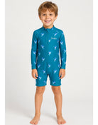 A young boy wearing a teal blue full-body swim suit with a white bird pattern, standing against a light gray studio background.