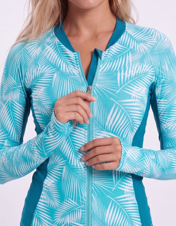 A woman shown from the front wearing a long-sleeve swim top with a turquoise and white palm leaf pattern and solid teal side panels, against a plain white studio background.