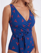 A blonde model wearing a blue one-piece swimsuit with a red lobster pattern and a tie waist, photographed against a plain white studio background close up