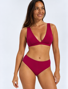 A woman modelling a two-piece bikini set in a plum colour, top plunge bikini and  bottom mid waist bikini, against a light grey studio background.