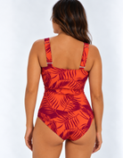 A woman shown from the back wearing aN eco one-piece swimsuit with a bold sunset palm tropical leaf pattern in bright orange and plum set against a plain light gray studio background.