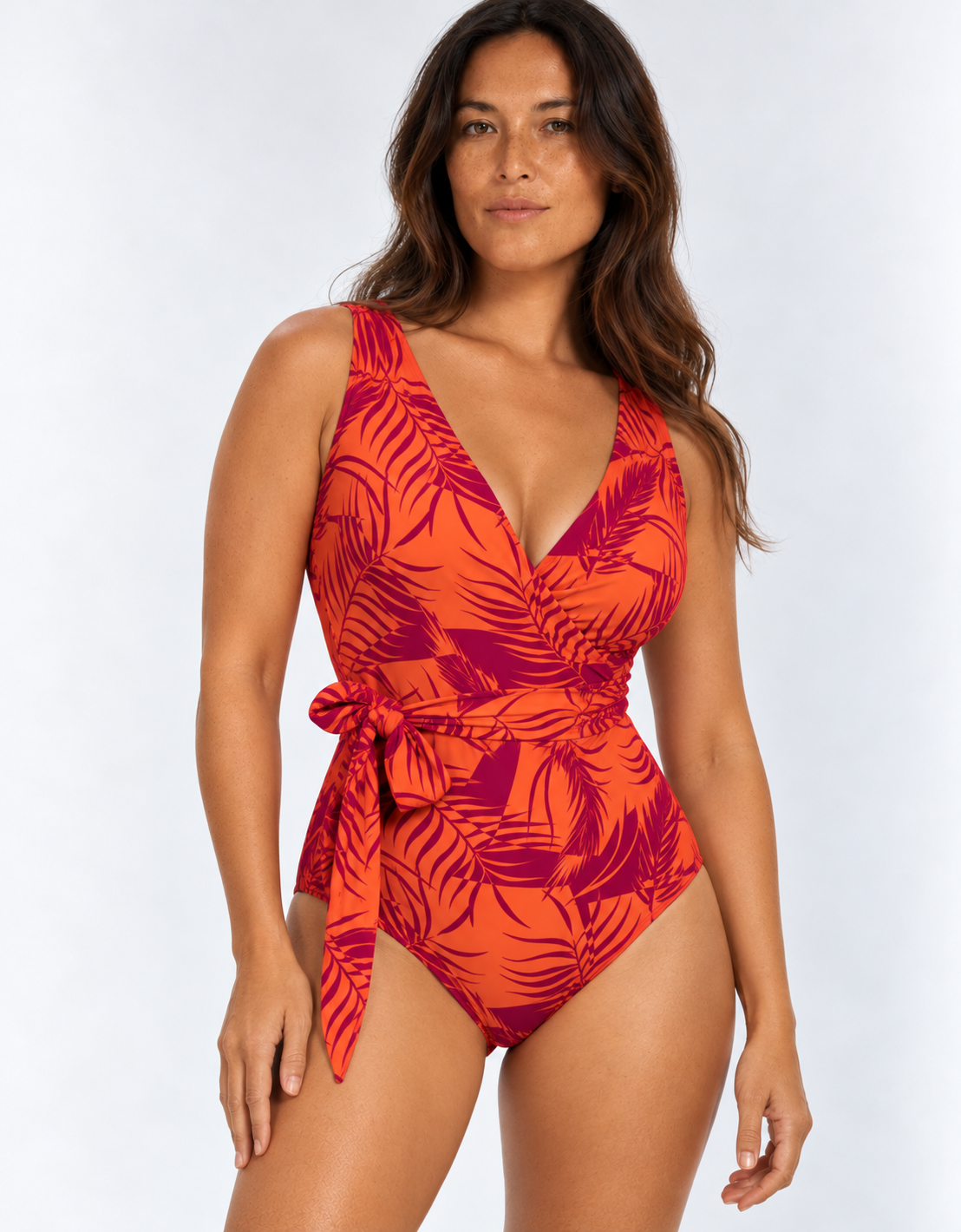 A woman modeling a one-piece swimsuit with a vibrant orange and dark red tropical leaf print, featuring a wrap-style V-neckline and a matching side tie, set against a plain light gray studio background.