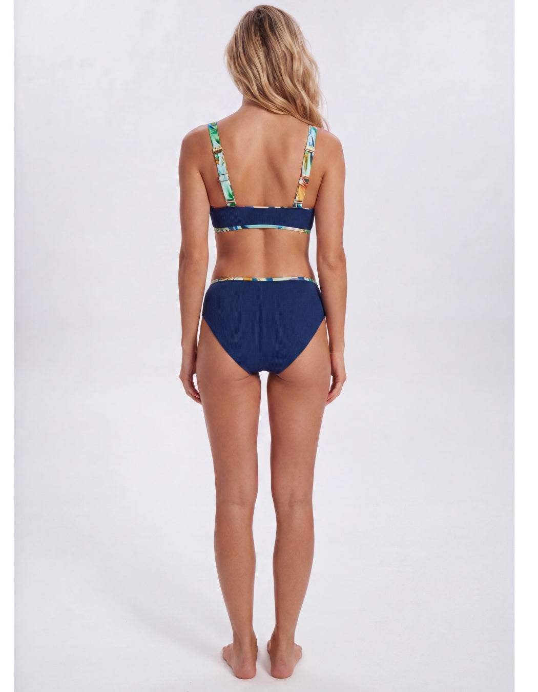 A woman shown from the back wearing a two-piece bikini. The bottom is solid navy blue, while the top has a tropical leaf print with green, yellow, and blue on a white base. Sandbar Swimwear