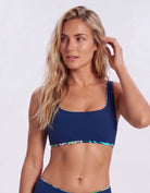 A blonde woman modeling a navy blue bikini top with a colorful patterned trim against a light gray studio background. Sandbar Swimwear