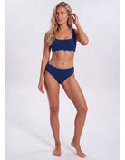 A woman modeling a two-piece navy blue bikini with a subtle multicolor patterned trim on the underbust band and waistband, photographed against a light gray studio background. Sandbar Swimwear