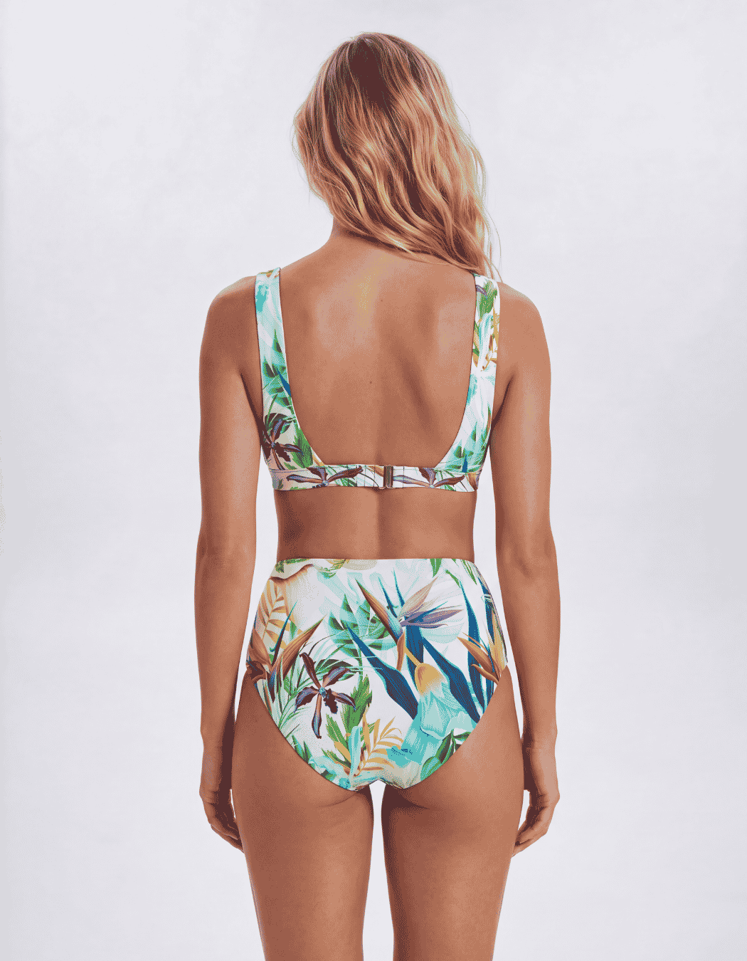 A woman shown from the back wearing a two-piece bikini with a tropical floral print. The swimsuit features a light blue, green, and white pattern with brown and orange accents, set against a plain light gray studio background. Sandbar Swimwear