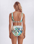 A woman shown from the back wearing a two-piece bikini with a tropical floral print. The swimsuit features a light blue, green, and white pattern with brown and orange accents, set against a plain light gray studio background. Sandbar Swimwear
