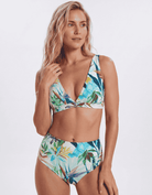 A woman modeling a two-piece bikini swimsuit with a tropical leaf print in shades of blue, green, and white, set against a plain light gray studio background. Sandbar Swimwear