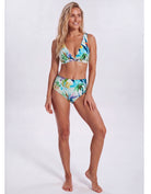 A woman modeling a two-piece bikini swimsuit with a tropical palm tree print in shades of blue, green, yellow, and white, set against a plain light gray studio background. Sandbar Swimwear