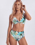 A woman modeling a two-piece bikini with a tropical leaf print in shades of green, blue, and white against a light gray studio background. Sandbar Swimwear