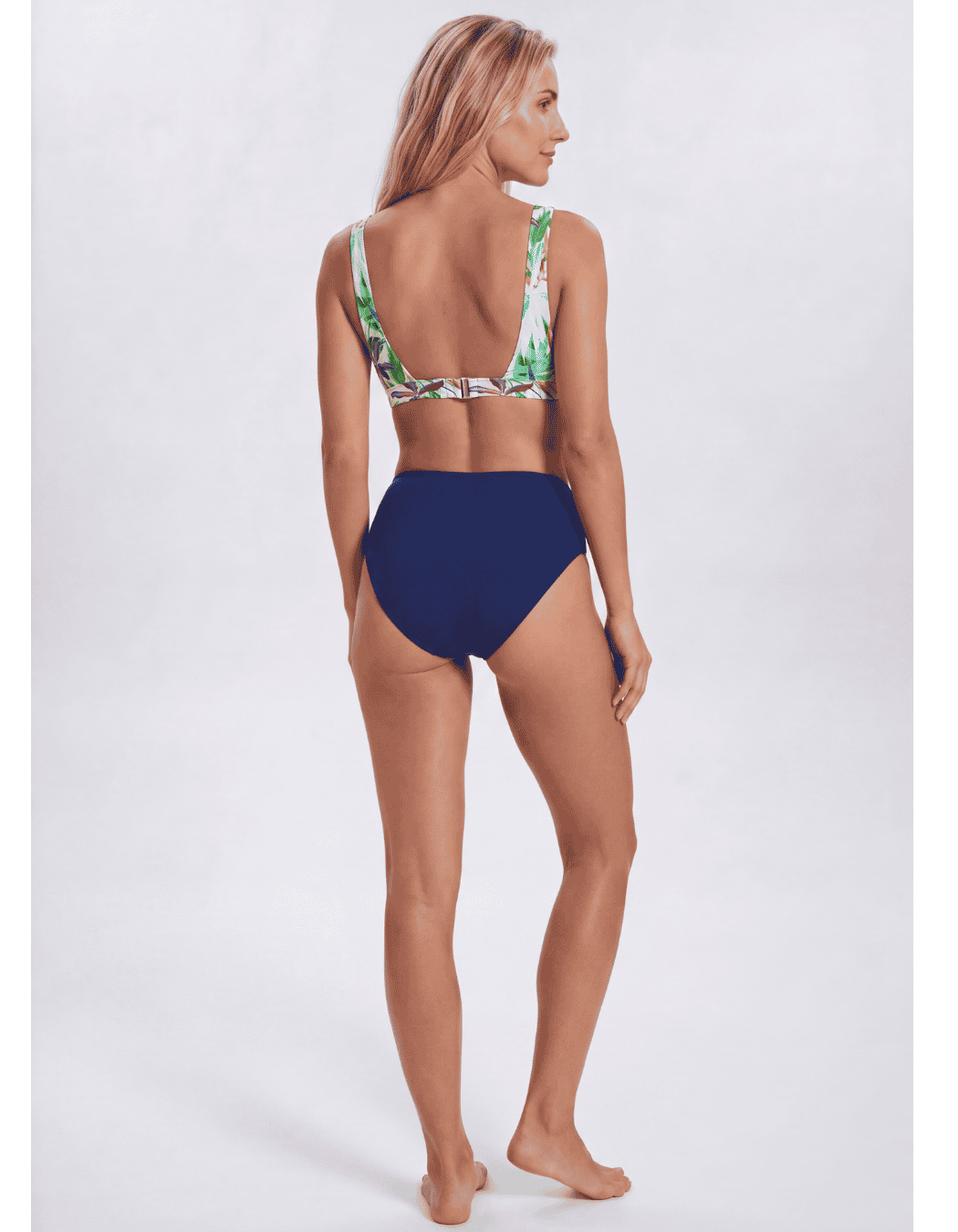 A woman modeling a two-piece bikini from the back, featuring a solid navy blue high-waisted bottom and a green tropical leaf print halter top, against a light gray studio background. Sandbar Swimwear