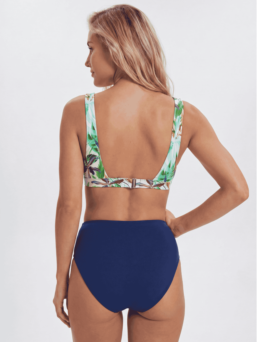 A woman shown from the back wearing a two-piece swimsuit with a navy blue high-waisted bottom and a tropical leaf print bikini top, against a light gray studio background. Sandbar Swimwear