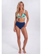 A woman modeling a two-piece bikini against a plain light gray studio background. The bikini top has a tropical floral print with green, blue, yellow, and white tones, while the high-waisted bottoms are solid navy blue. Sandbar Swimwear