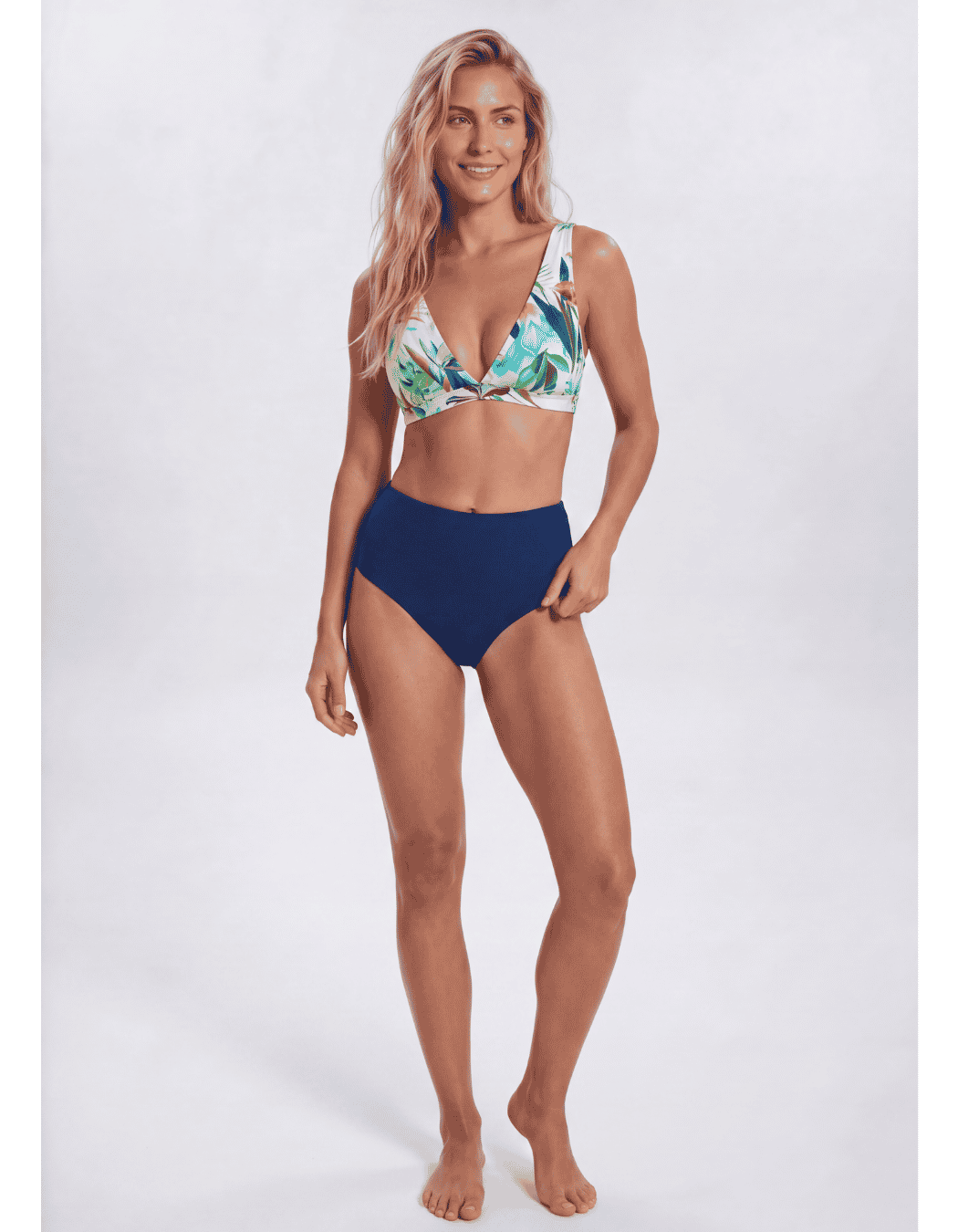 A woman modeling a two-piece bikini against a plain light gray studio background. The bikini top has a tropical leaf print in green, blue, and white, while the high-waisted bottoms are solid navy blue. Sandbar Swimwear