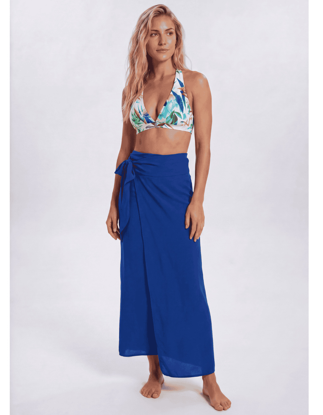 A woman modeling a royal blue wrap sarong skirt paired with a tropical print bikini top, standing against a light gray studio background. Sandbar Swimwear