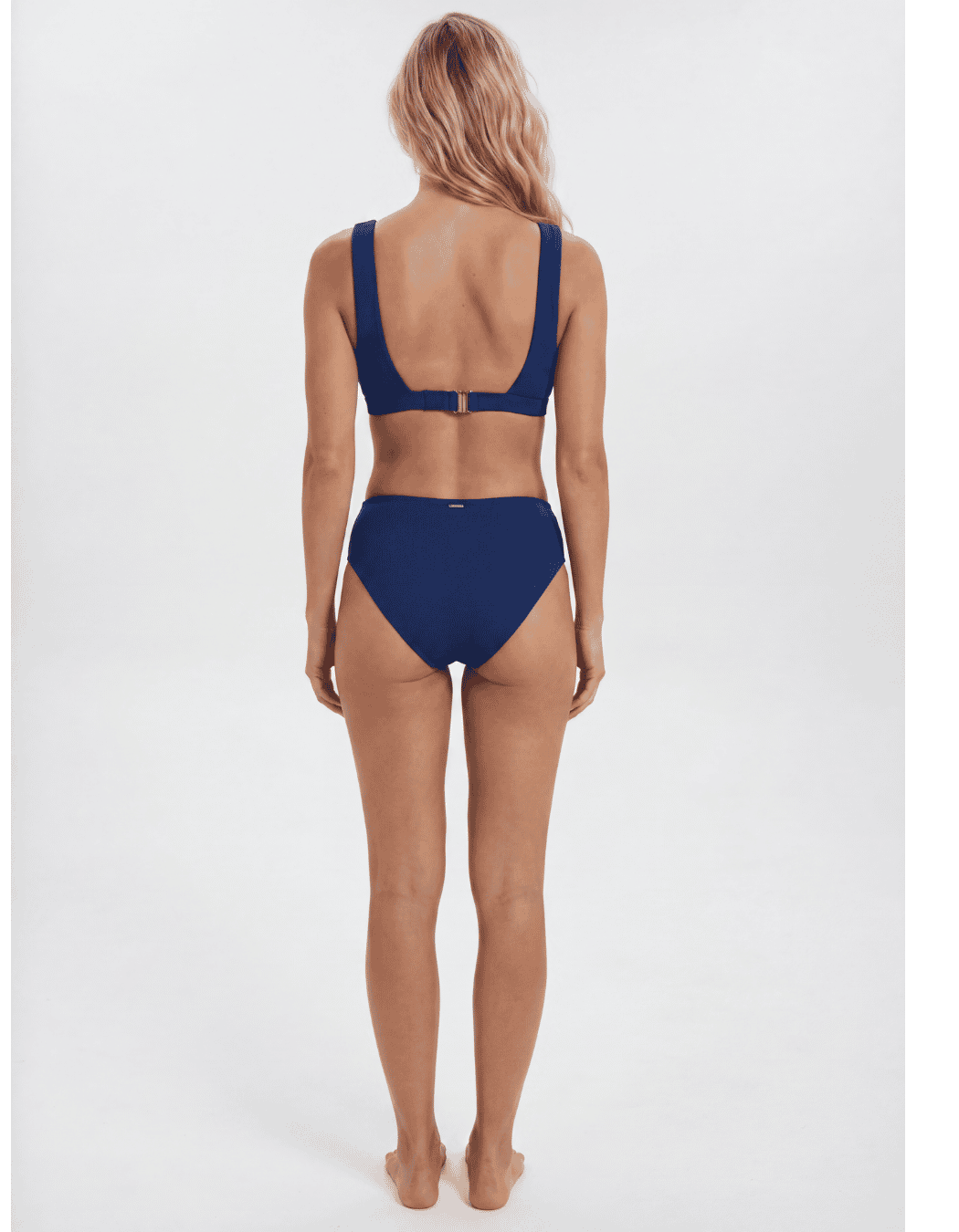 A woman viewed from the back wearing a navy blue two-piece bikini against a plain light gray studio background. Sandbar Swimwear