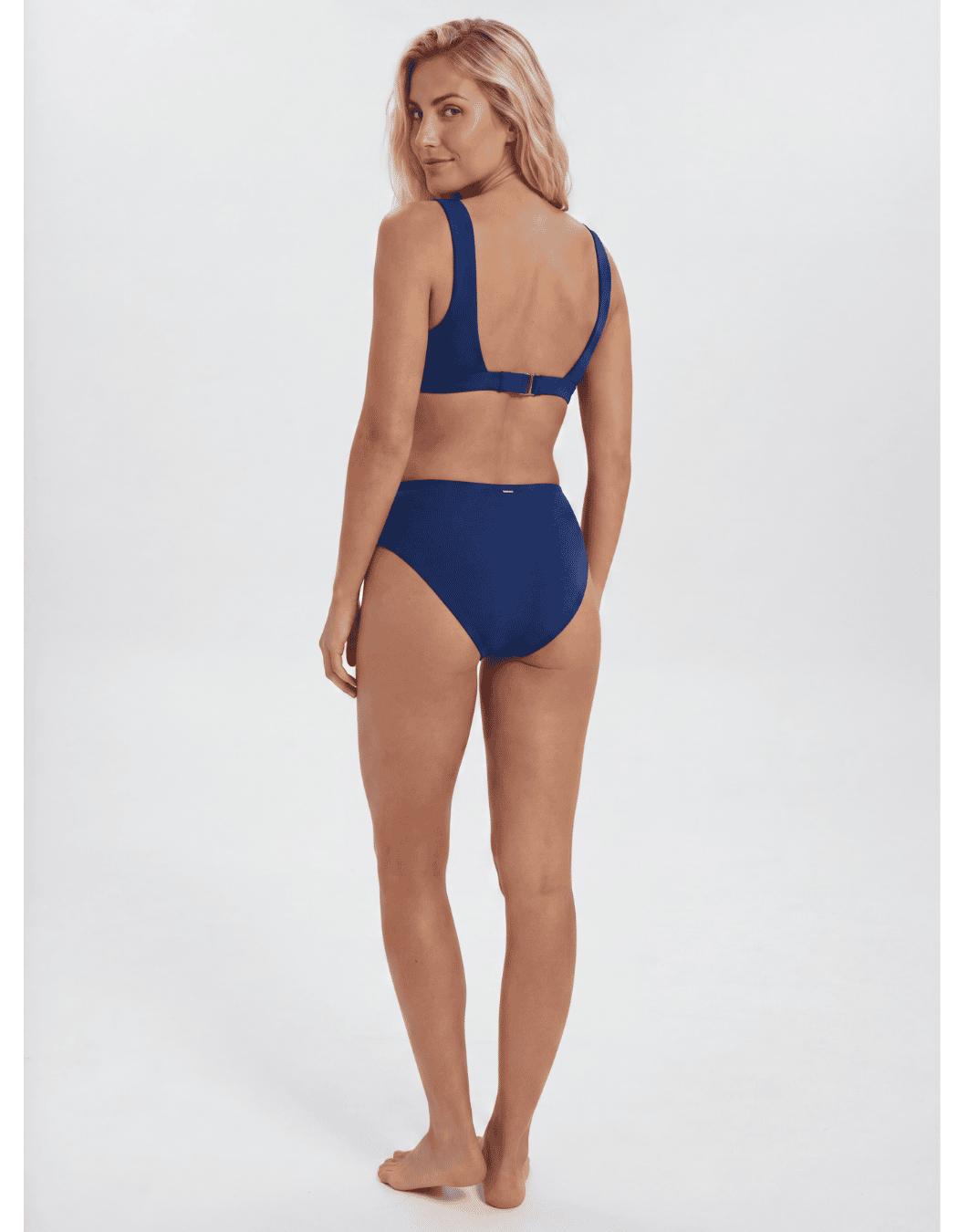 A woman modeling a two-piece navy blue bikini from the back, standing against a light gray studio background. Sandbar Swimwear