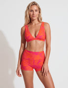 A woman modeling a two-piece bikini set with a vibrant neon orange and pink tropical leaf pattern against a plain white studio background.