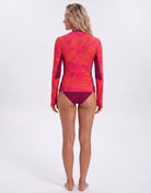 A woman stands with her back to the camera wearing a long-sleeve bikini top and matching bottoms. The top features a bright pink and orange tropical leaf pattern, with solid dark magenta side panels and a solid dark magenta bottom.