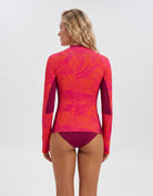 A woman shown from the back wearing a long-sleeve swim rash guard with a pink and orange tropical leaf pattern and solid magenta side panels, paired with matching magenta swim bottoms, against a light gray studio background.