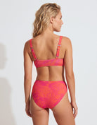 A woman modeling a two-piece bikini from the back, featuring a vibrant pink and orange tropical leaf pattern, set against a plain light gray studio background.