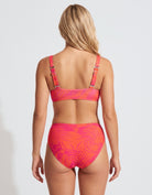 A woman shown from the back wearing a two-piece bikini with a bright pink and orange tropical leaf pattern, set against a plain light gray studio background.