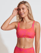 A woman modeling a two-piece bikini set with a vibrant pink and orange tropical leaf pattern against a plain light gray studio background.